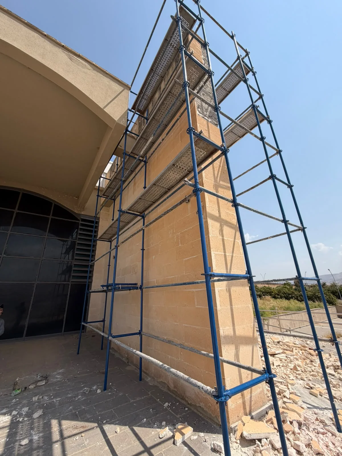 ASL YAPI — Taş kaplı yapı köşesinde mavi flanşlı dikmeler, yatay bağlantılar ve delikli metal platformlar